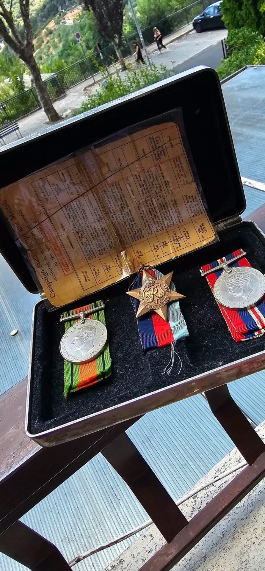 Open presentation case displaying three medals with striped ribbons beside an aged document, set outdoors on a railing ledge with trees and a hillside in the background.