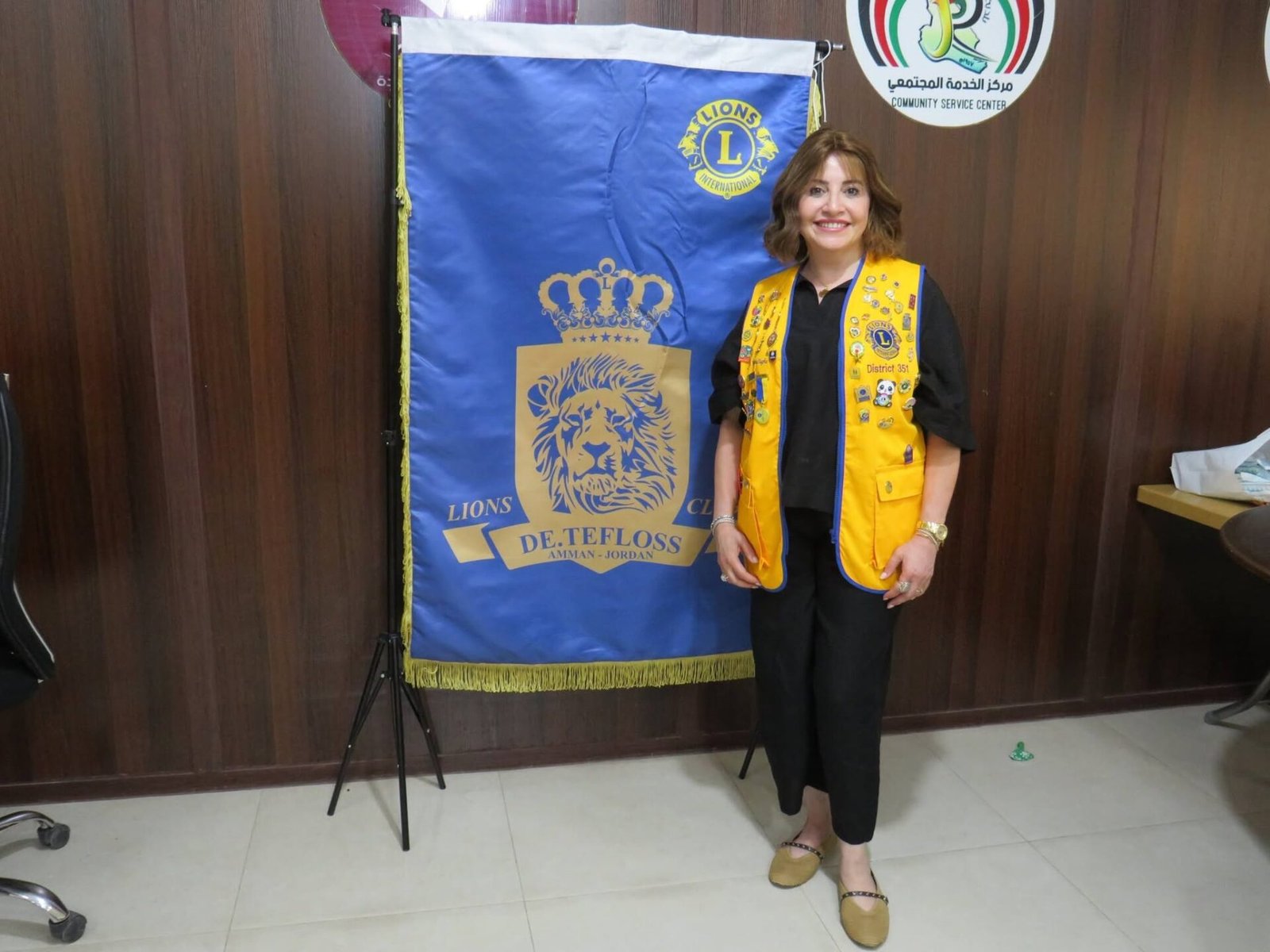 768230425_122373551576016148_4442548185917321252_n - عروبة الاخباري | Oroba News Smiling woman in a yellow Lions vest stands beside a tall blue Lions banner with a crowned lion emblem indoors.