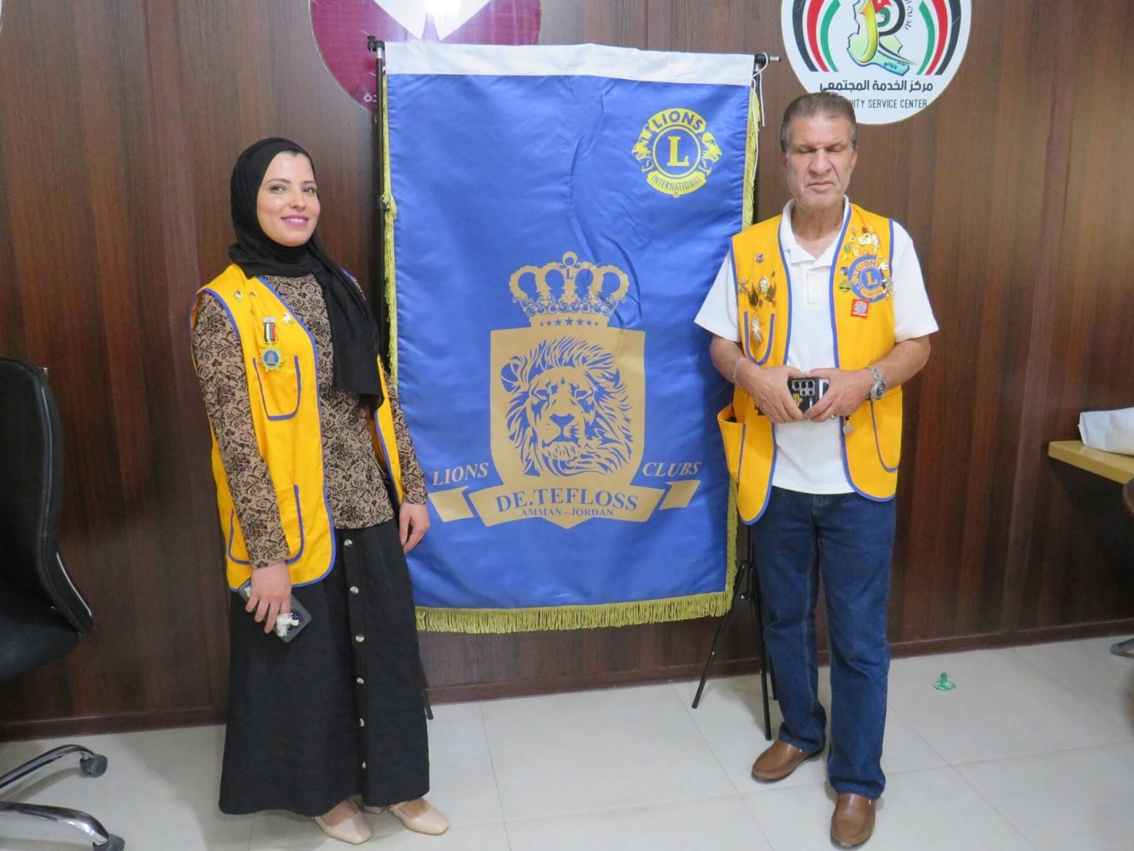 768347832_122373551666016148_1915443381578010712_n - عروبة الاخباري | Oroba News Two people in yellow Lions Club vests stand beside a blue Lions banner with a gold emblem in a room with wood-paneled walls and a round logo on the wall behind them.