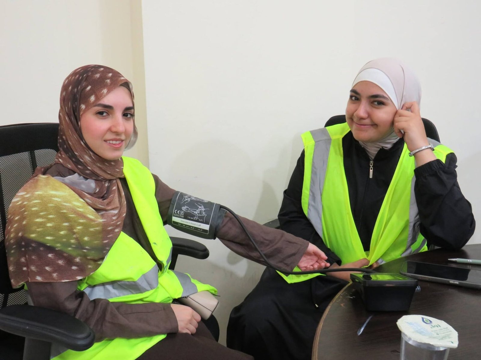 768408798_122373550784016148_556111675642221116_n - عروبة الاخباري | Oroba News Two women wearing hijabs and neon safety vests sit at a desk; one is taking the other's blood pressure with a cuff on the arm.