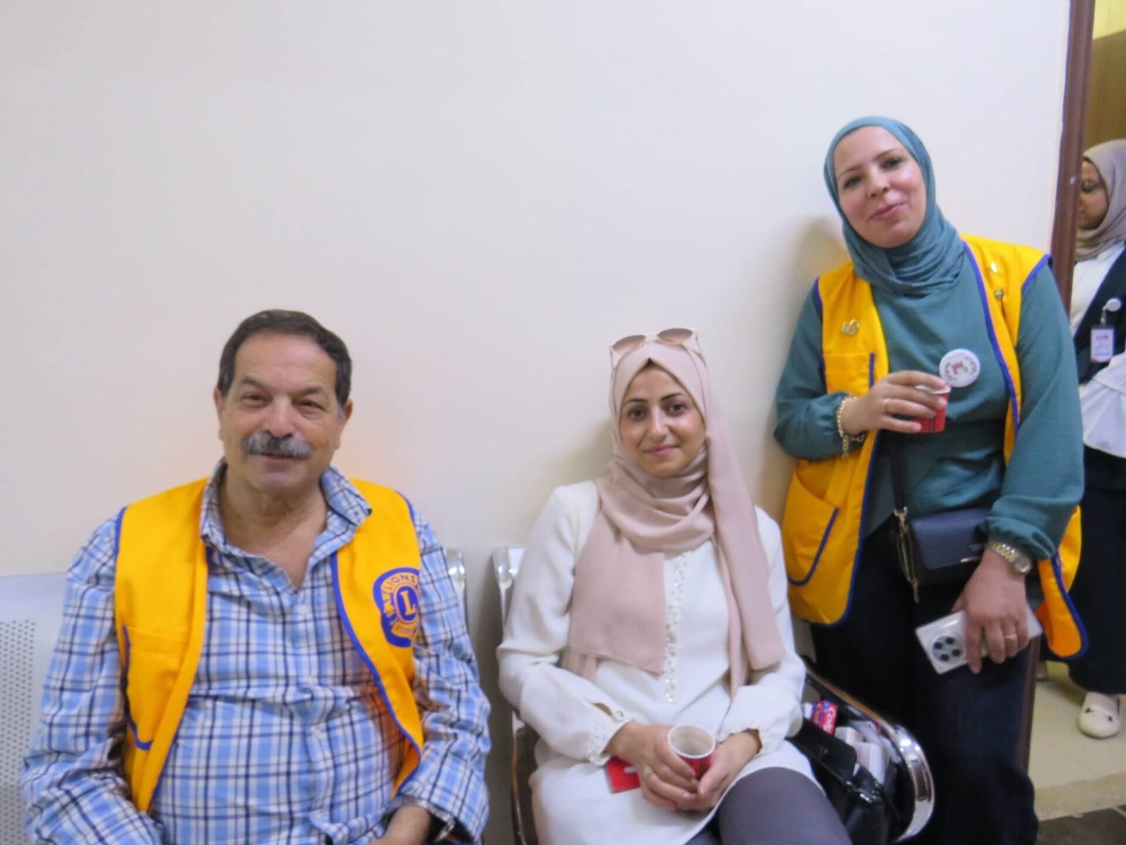 768452813_122373550976016148_8185100436079377268_n - عروبة الاخباري | Oroba News Group of four people indoors posing for a photo; two women in hijabs wearing yellow vests, one man in a yellow vest and plaid shirt, and a woman in white seated in front holding a red cup (partial view of a fourth person in background).