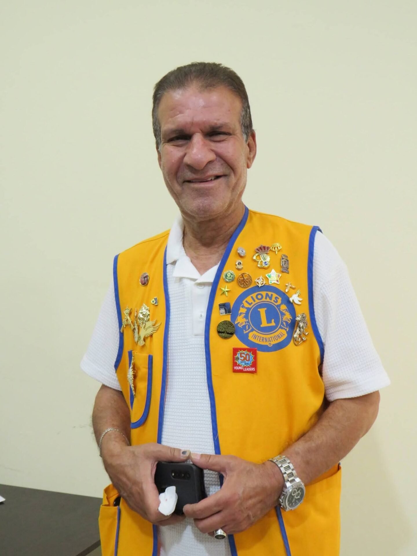768529188_122373551618016148_1260731554854396972_n - عروبة الاخباري | Oroba News Smiling man wearing a yellow Lions Club vest with pins, holding a phone indoors.