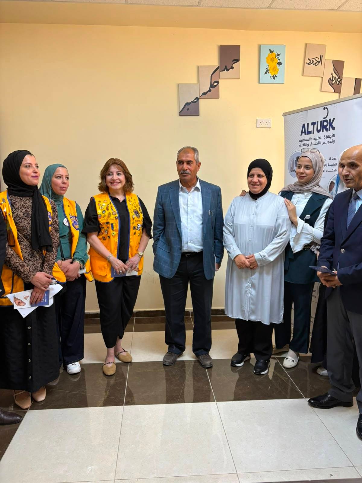 768555353_122373550232016148_1241572533209527764_n - عروبة الاخباري | Oroba News Group of adults posing for a photo indoors; several wear yellow vests, others in smart casual clothing, near a banner.