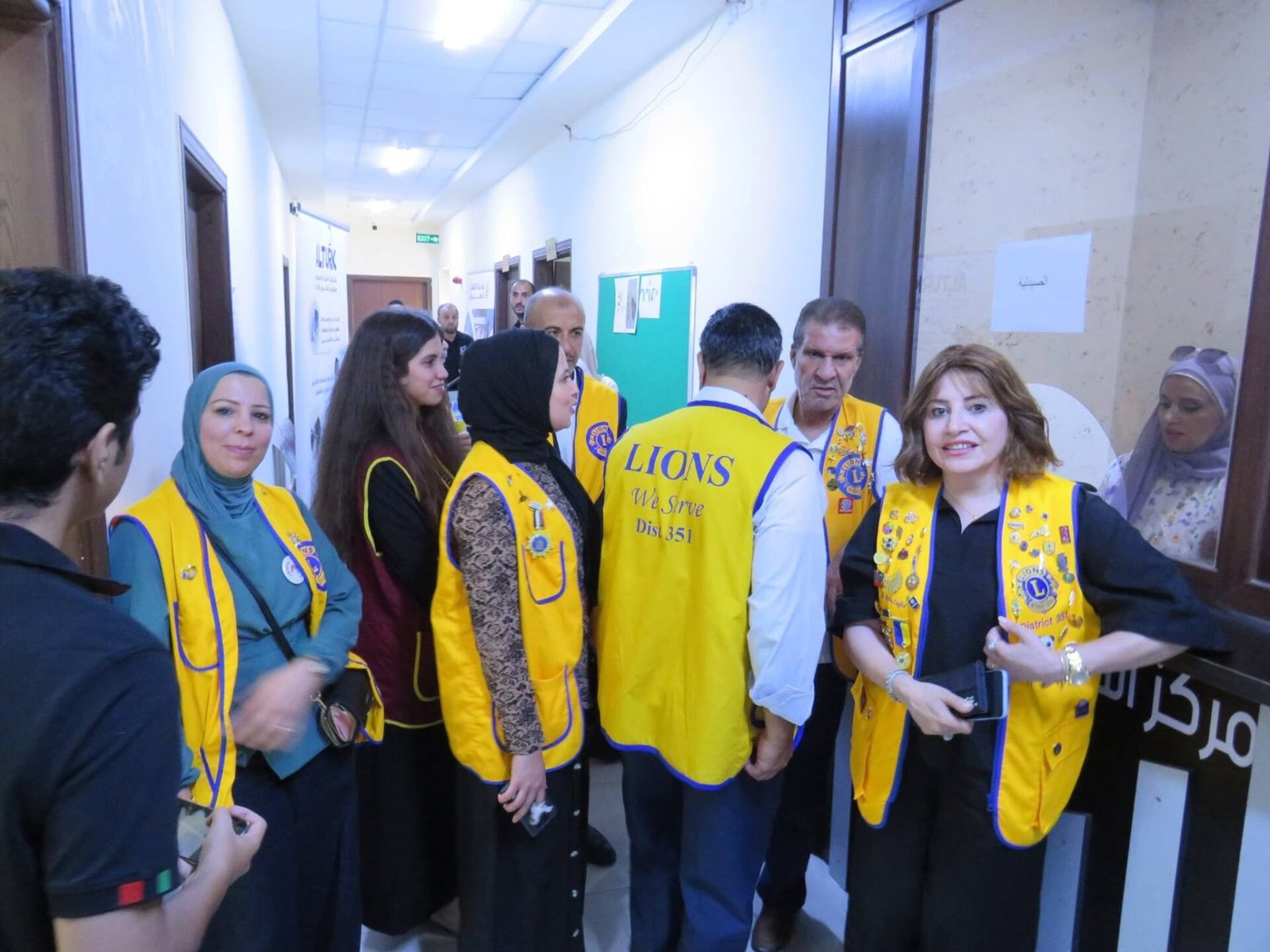 769051724_122373551018016148_24551599257602963_n - عروبة الاخباري | Oroba News Group of Lions Club volunteers in yellow vests standing in a hallway, conversing with each other.