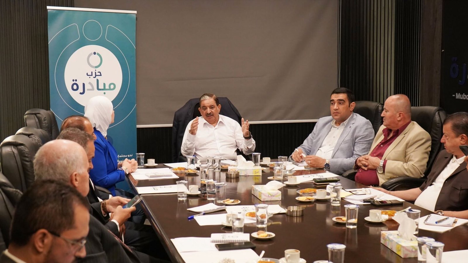 769131213_122145932373082282_3743791036514151579_n - عروبة الاخباري | Oroba News Group of professionals around a long conference table during a formal meeting, with a blue Arabic banner on the left.