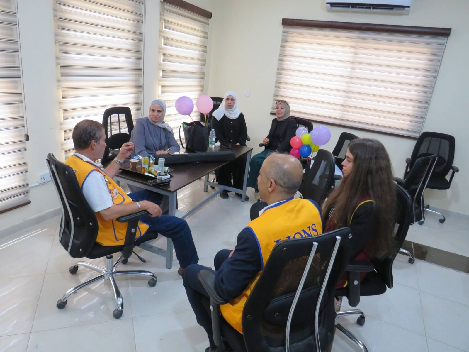 769144593_122373550742016148_3001531090302975831_n - عروبة الاخباري | Oroba News Group of six adults in an office meeting room, some wearing yellow vests, sitting around a table with balloons and drinks.