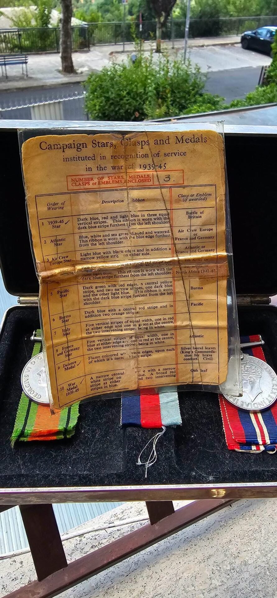 Display of WWII campaign medals with colorful ribbons and an aged document titled 'Campaign Stars, Clasps and Medals' on a bench outdoors