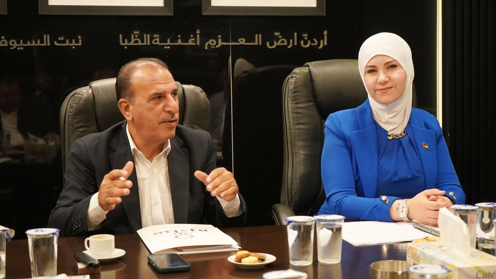 769182814_122145932361082282_3886499259605964906_n - عروبة الاخباري | Oroba News Two professionals seated at a conference table; man in a dark suit speaks while gesturing, woman in a royal blue blazer and white hijab listens beside him, with Arabic script on the dark wall behind them.