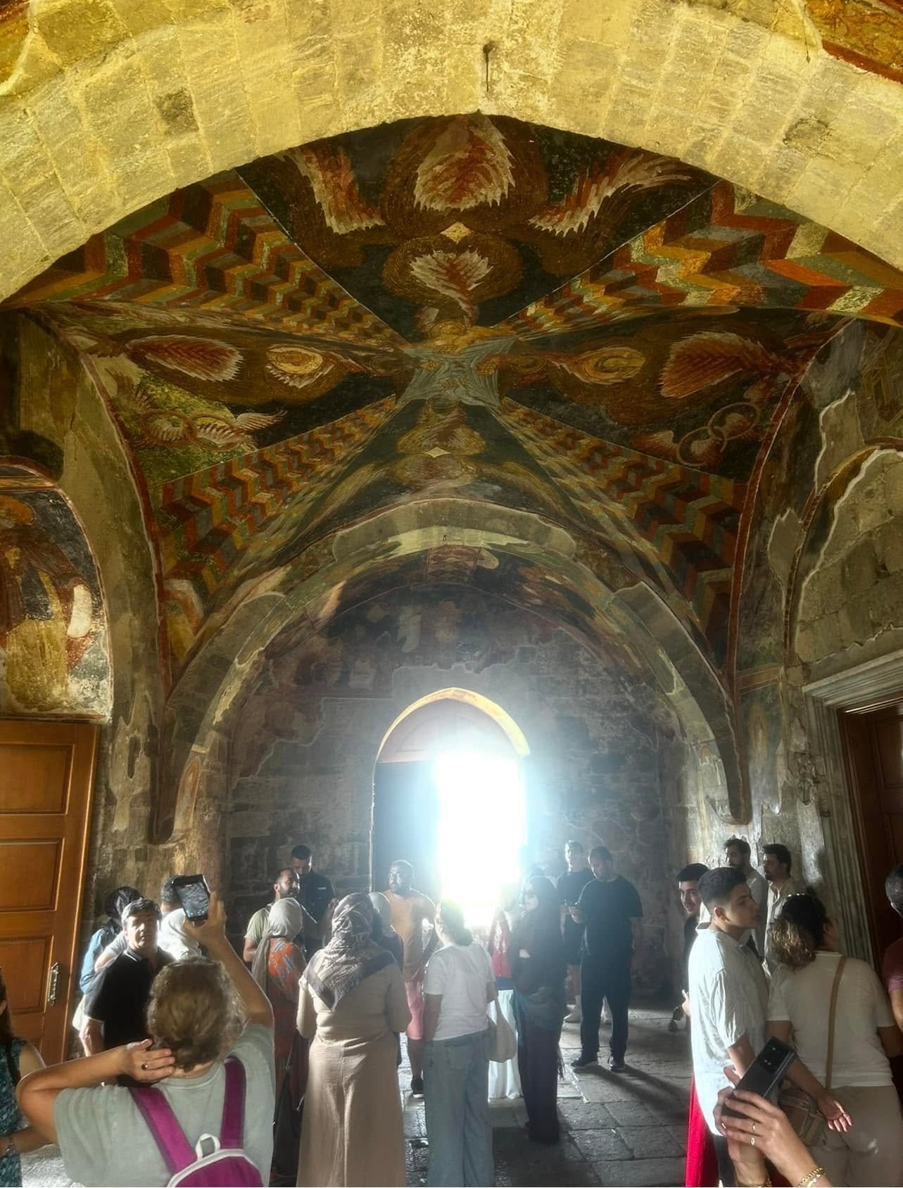 Screenshot - عروبة الاخباري | Oroba News Interior view of a stone arch ceiling covered in colorful frescoes and geometric patterns, with a crowd of people standing beneath the arches near a bright doorway.