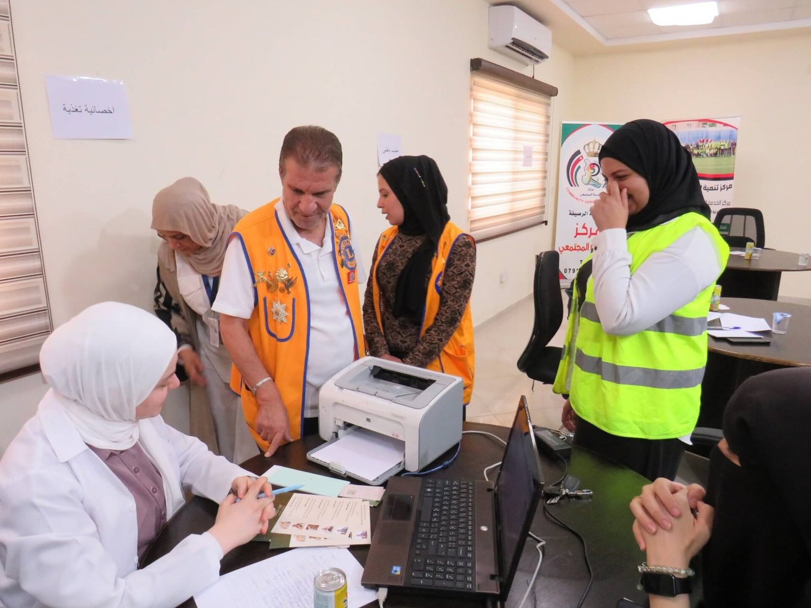 771024910_122373551168016148_3168223033465540414_n - عروبة الاخباري | Oroba News Staff and volunteers assist at a registration desk with a printer, laptops, and papers on the table in a community center setting.