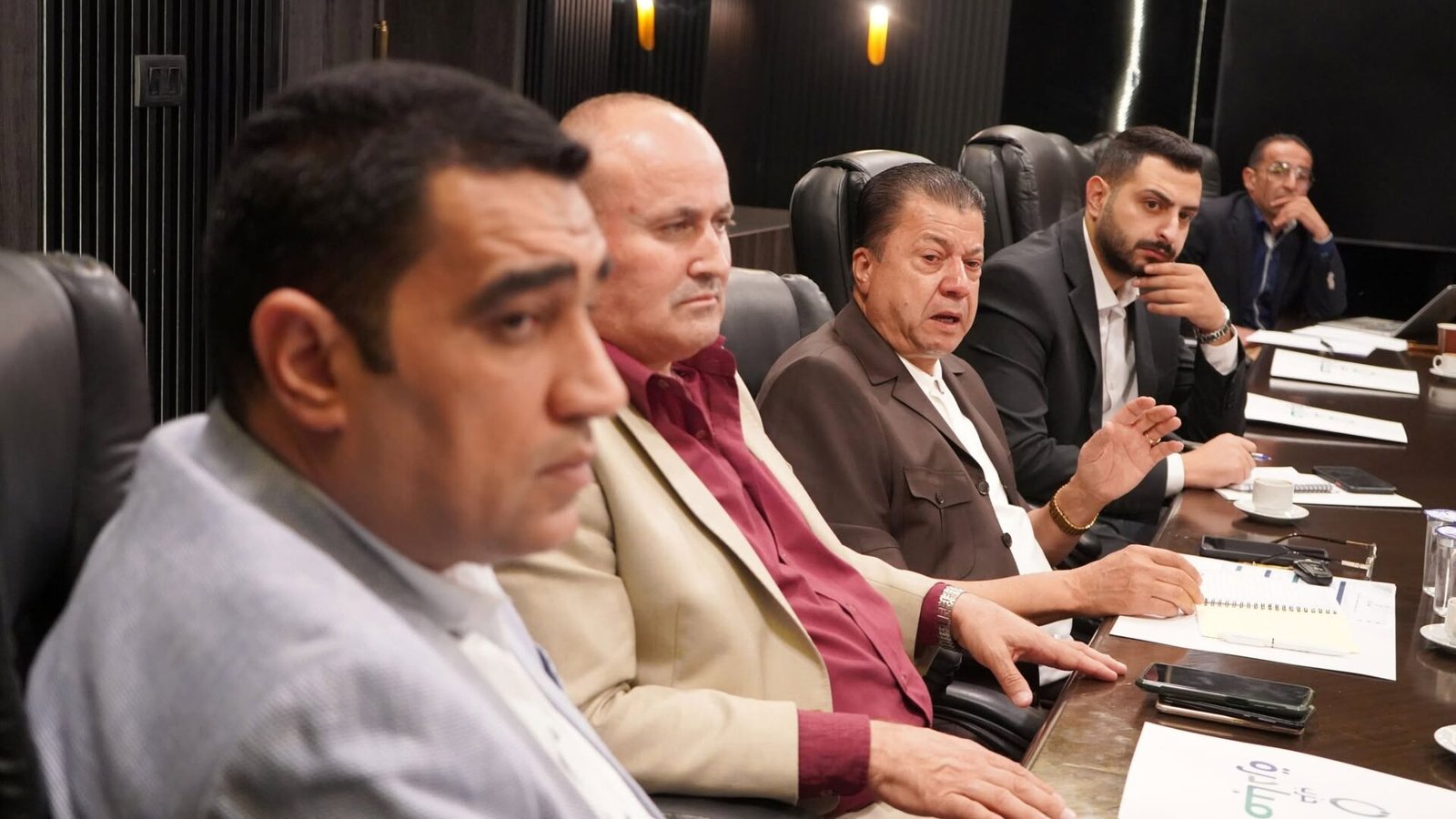 771795763_122145932163082282_2211441511116009699_n - عروبة الاخباري | Oroba News Five men in business attire sit along a conference table, reviewing documents in a dim, upscale boardroom.