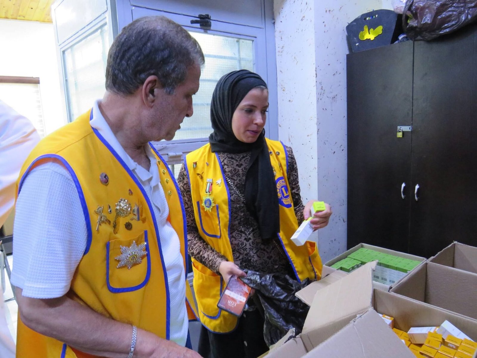 772138435_122373550874016148_966283525629078955_n - عروبة الاخباري | Oroba News Two volunteers in yellow vests inspect boxes of small yellow and green items on a table in a storage room.