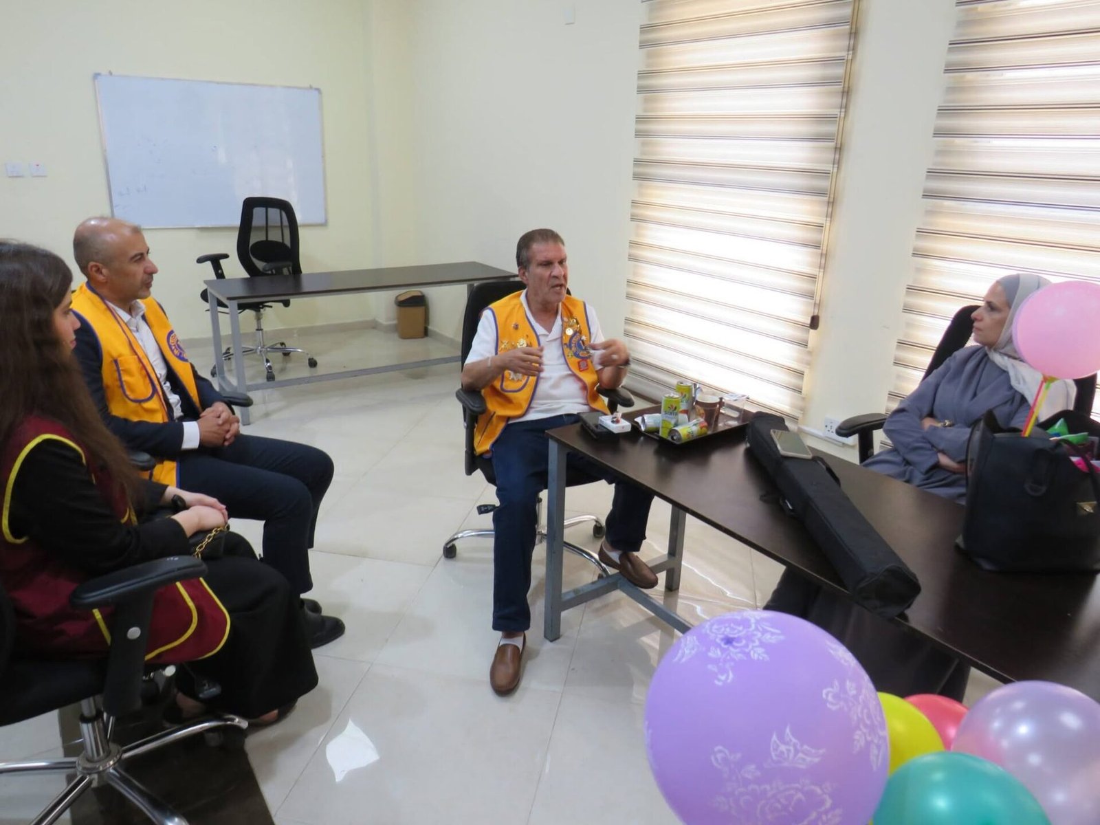 772632565_122373550508016148_4418866889813215291_n - عروبة الاخباري | Oroba News Group of people wearing yellow vests sitting in a circle, listening to a man speaking in a meeting room with balloons in the foreground and a whiteboard in the background.