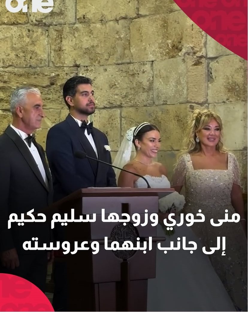 Bride and groom in formal attire stand at a podium with two well-dressed guests beside them during a wedding ceremony, against a stone wall, with a large white Arabic caption across the bottom.