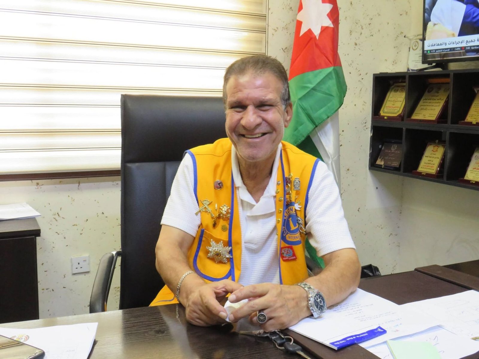 773093639_122374744226016148_4503780712018165182_n - عروبة الاخباري | Oroba News Smiling man in an orange Lions Club vest sits at a desk with papers, a watch, and jewelry, in an office with a Jordan flag in the background.