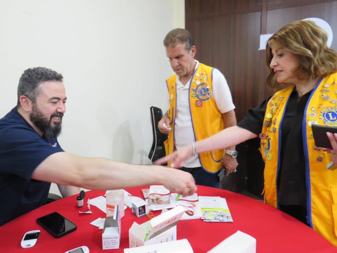 773241923_122374743962016148_312833034159442672_n - عروبة الاخباري | Oroba News Bearded man in a navy shirt shakes hands with two Lions Club volunteers in yellow vests at a table with brochures on display.