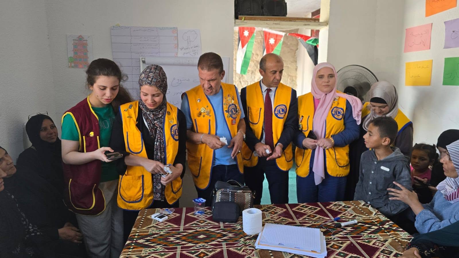 773544640_122374744274016148_7677395405789970611_n - عروبة الاخباري | Oroba News Group of Lions Club volunteers in yellow vests handing out supplies to women and children in a community room.