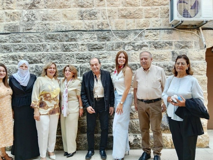 Eight adults stand in a row outside a textured stone wall, dressed in smart casual to semi-formal attire.