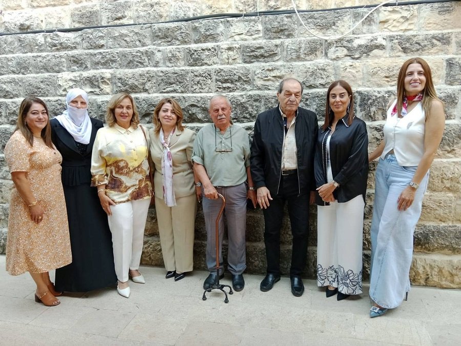 Nine adults pose in a row against a textured stone wall, smiling, with an elderly man using a cane in the center.