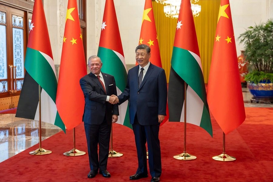 Two men in suits shake hands on a red carpet, with Chinese and Jordanian flags behind them in a formal hall.