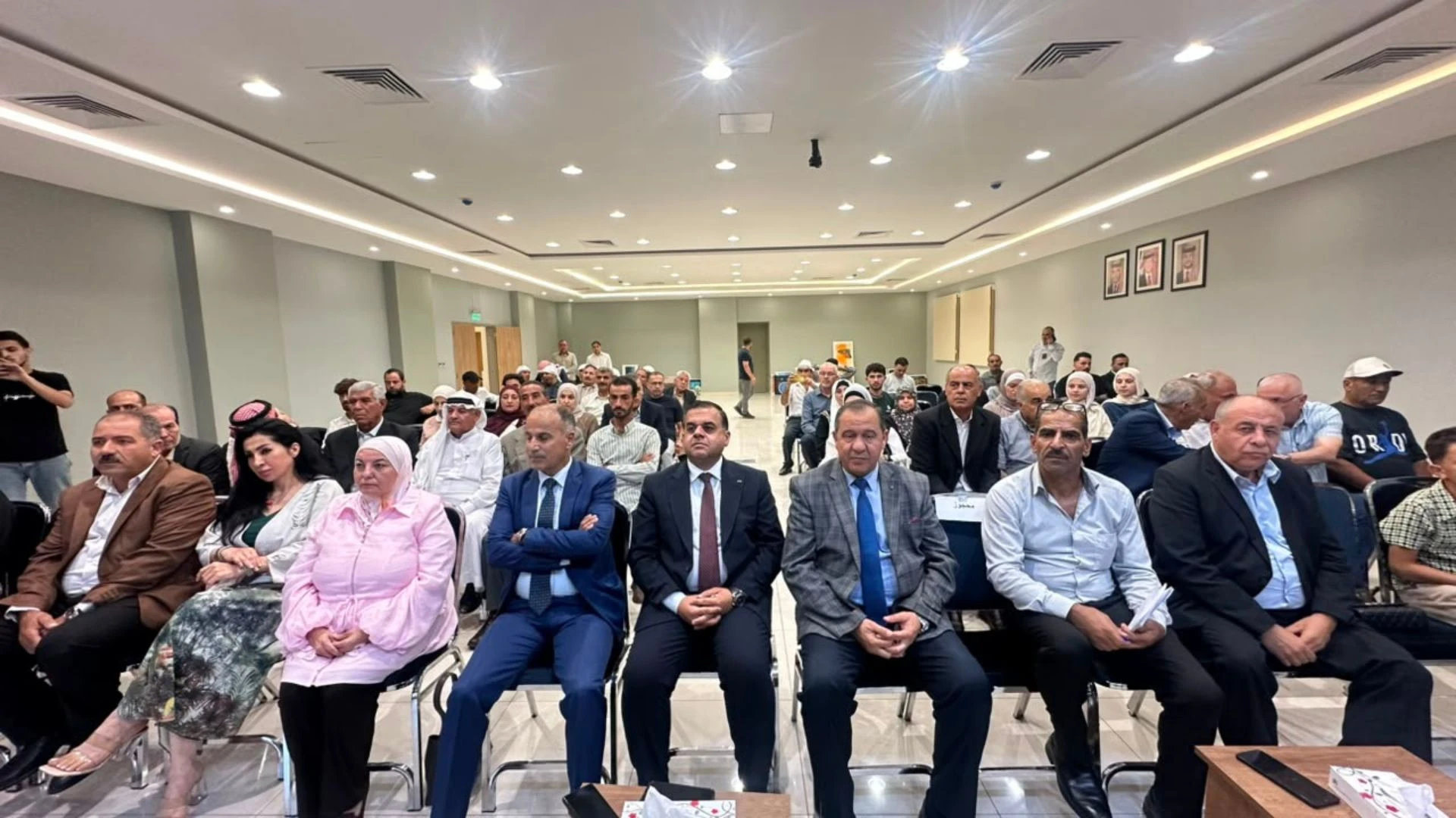 78249c31-3dbd-4475-814b-bf30915fcf2f - عروبة الاخباري | Oroba News A large meeting room filled with men and women in formal attire seated in rows for a conference or presentation in a bright, modern hall.