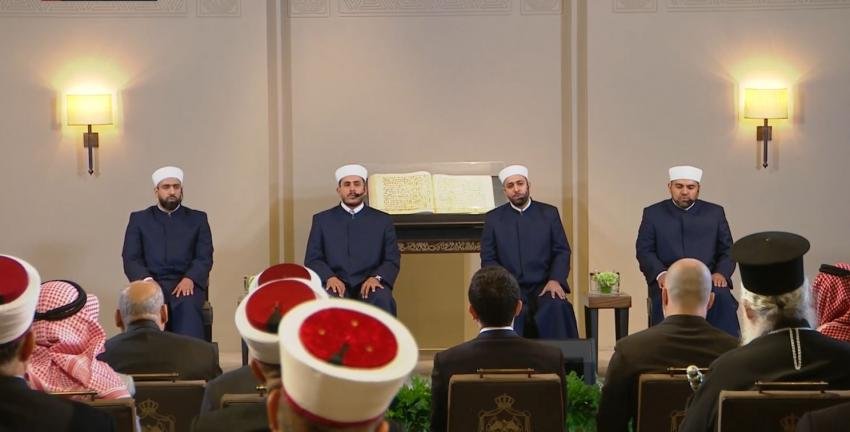 Four men in dark blue robes and white caps sit in a row on a stage with an open Quran on a stand, addressing a seated audience at a formal religious gathering.