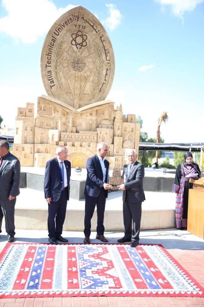 D304uY8A6sOmoYGKwrKjzXeI0UHNXfsLCUfiyPiZ - عروبة الاخباري | Oroba News Three men in suits present a trophy in front of a large stone monument at Tafila Technical University under a clear sky.