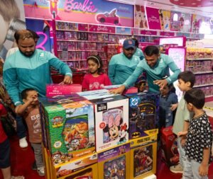Deliveroo x Toys R Us - 15 - عروبة الاخباري | Oroba News Staff in teal uniforms assist children at a crowded toy counter with colorful boxed toys in a Barbie-themed shop outside the camera's view
