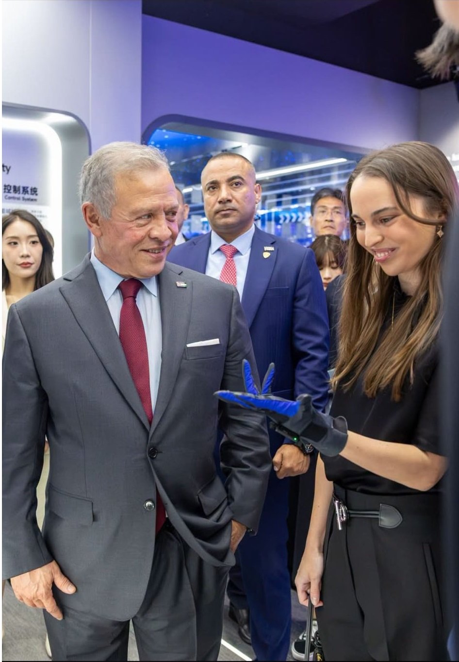 HQAH7O7WAAEztM_ - عروبة الاخباري | Oroba News Smiling man in a gray suit and red tie chats with a woman in black, who wears a blue glove and shows him something at a tech exhibition.