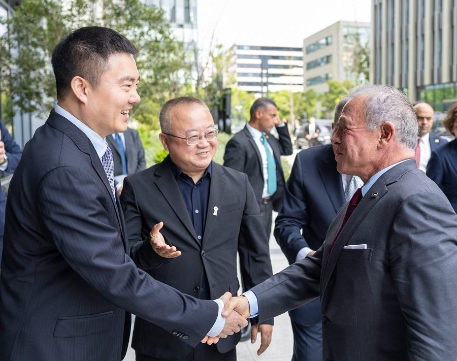 HQAH7O9XcAAFQqz - عروبة الاخباري | Oroba News Two men in suits shake hands in an outdoor business gathering, smiling, with other professionals in the background.