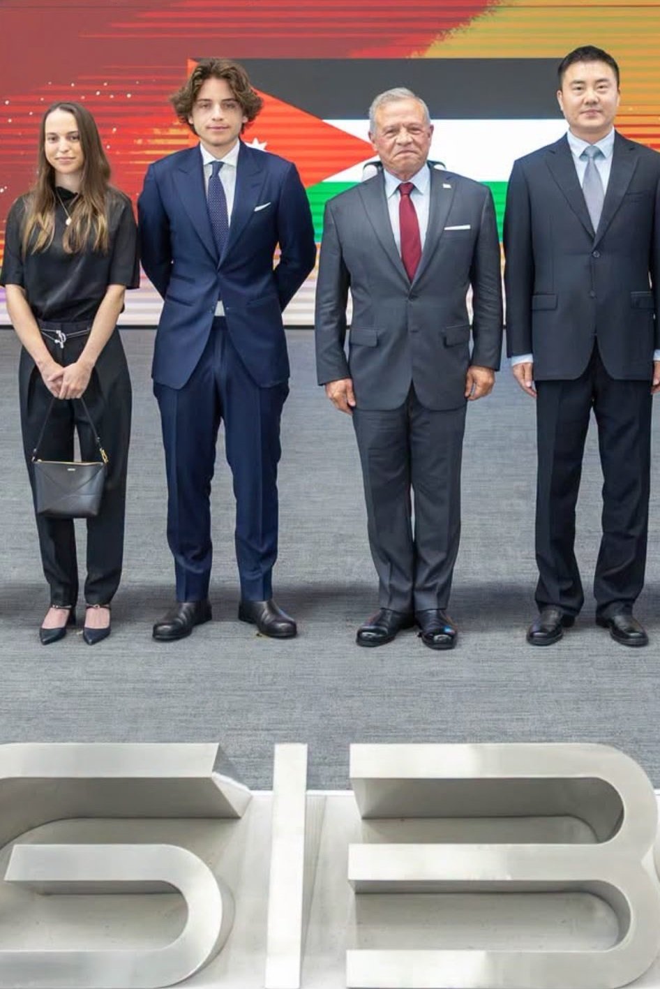 HQAH7O_XMAAJlUf - عروبة الاخباري | Oroba News Four formally dressed people posing for a group photo at a formal event, with a flag backdrop behind them.