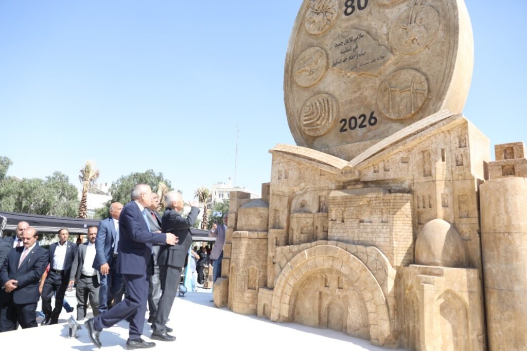 PIjDJZIvu4RWG5xMmssXPf9J0vZeaEDGoQvzNqTL - عروبة الاخباري | Oroba News Dignitaries in suits walk toward a large sandstone monument with circular reliefs and the year 2026 inscribed on it.