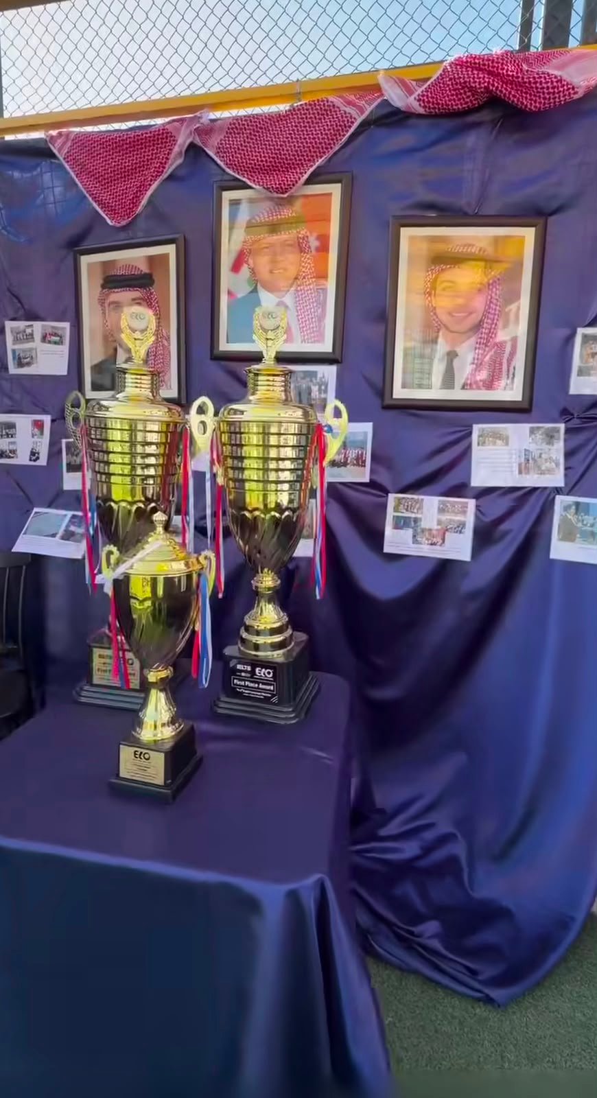 WhatsApp Image 2026-08-01 at 33333 PM 2 Three gold trophies on a purple table in front of a photo wall and portrait frames framed by a purple backdrop.