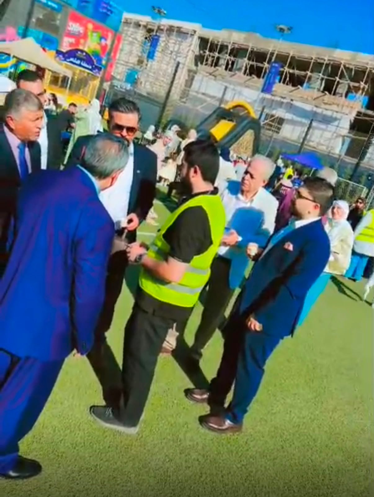 WhatsApp Image 2026-08-01 at 33334 PM 2 Group of men in suits conversing with a worker in a bright safety vest on a green sports field near a playground under construction.