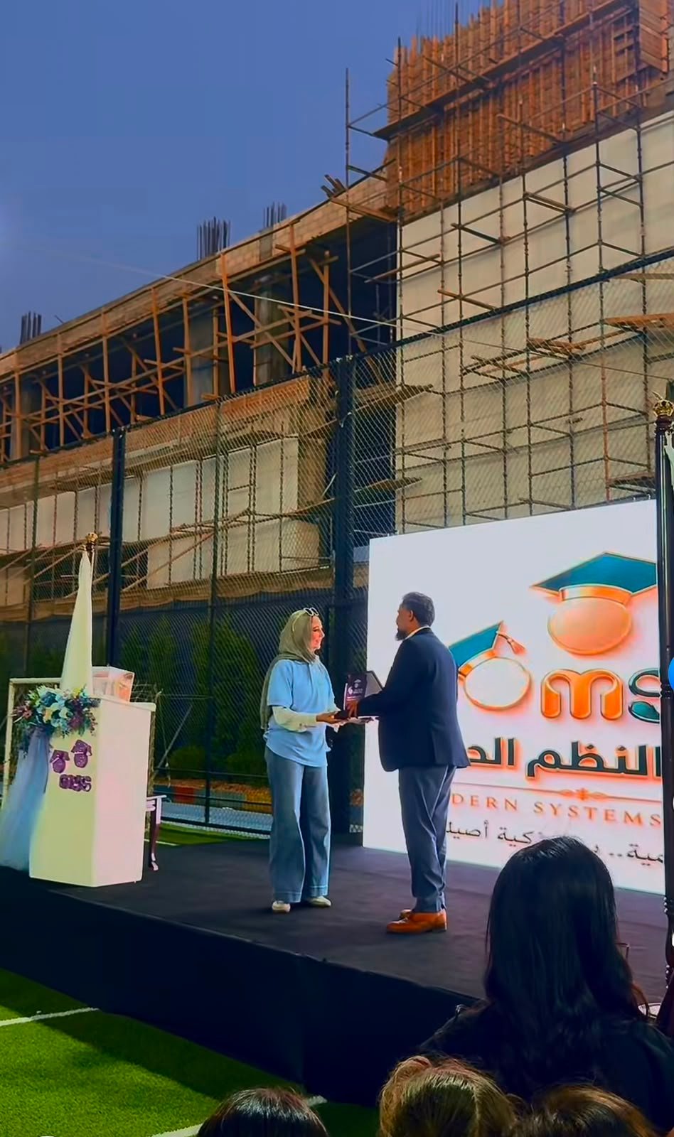 WhatsApp Image 2026-08-01 at 33334 PM 5 Woman in a blue hijab and matching outfit receives an award on a stage from a man in a suit, against a construction site backdrop and a large screen with graduation graphics.