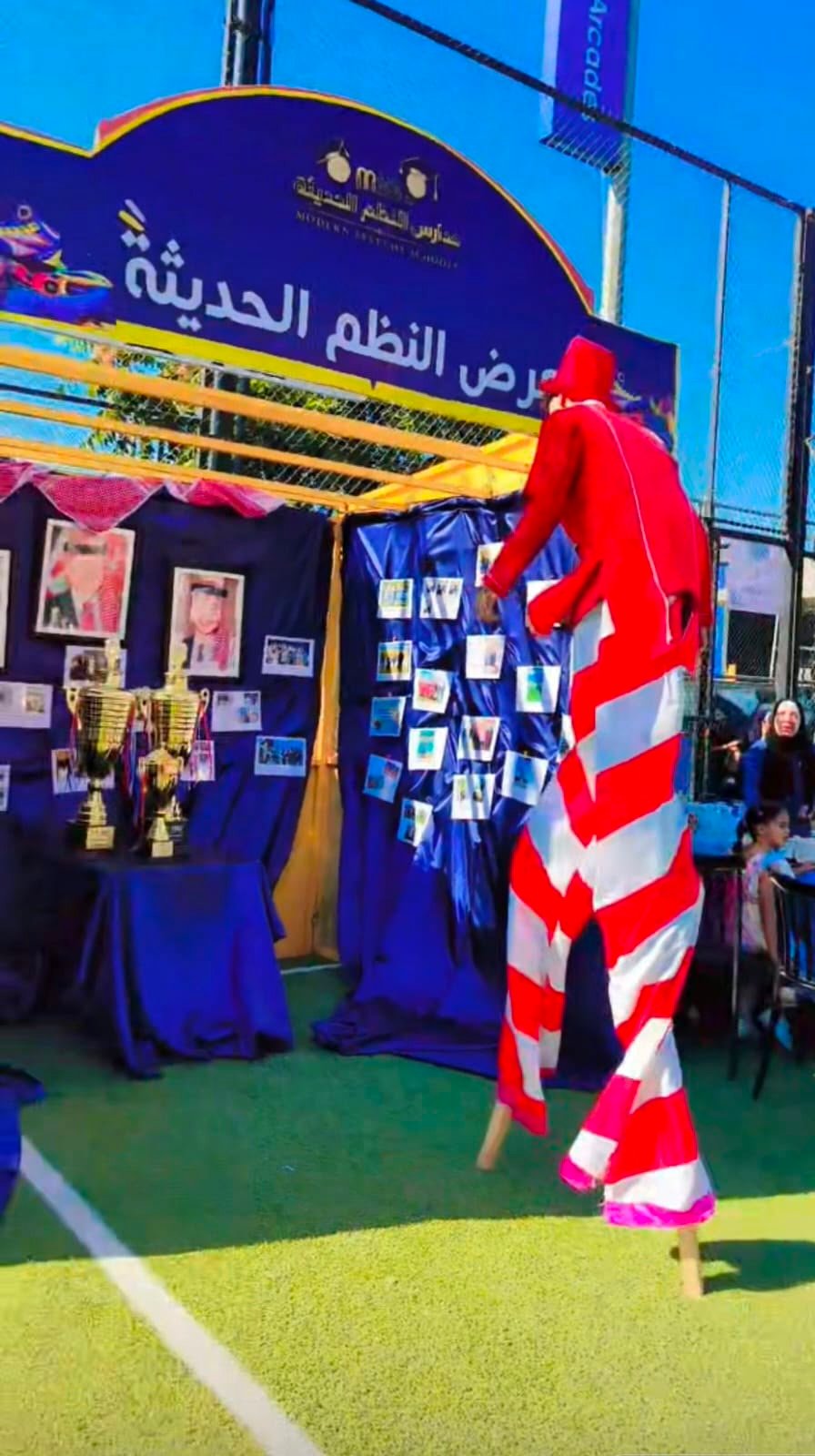WhatsApp Image 2026-08-01 at 33334 PM 6 Stilt performer in a red-and-white striped suit beside a festival booth with photos and trophies outside.