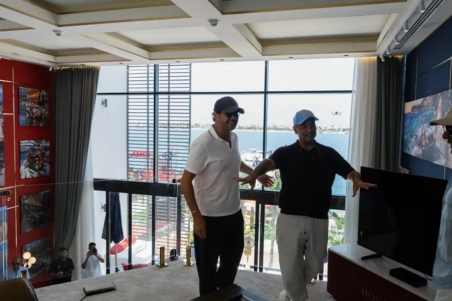 WhatsApp Image 2026-08-01 at 80018 PM1 Two men in caps stand in a bright, modern room by large windows overlooking the water, gesturing toward a TV.