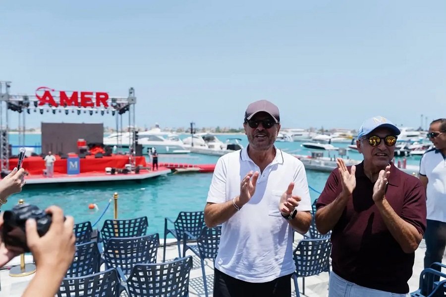 WhatsApp Image 2026-08-01 at 80018 PM2 Two men in casual polo shirts clap at a waterfront event, with a stage and boats in the harbor behind them.