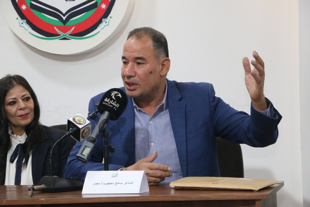 WhatsApp Image 2026-08-04 at 111734 AM Man in a blue suit speaking at a press conference, gesturing with his right hand while microphones are in front; woman sits beside him and a crest on the wall is visible.