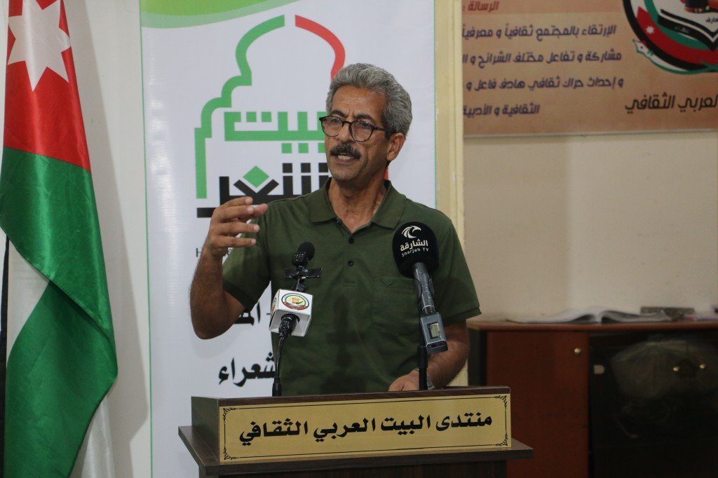 WhatsApp Image 2026-08-04 at 111736 AM Man with gray hair and glasses speaks at a podium with two microphones, a flag on the left and a green-logo banner behind him.