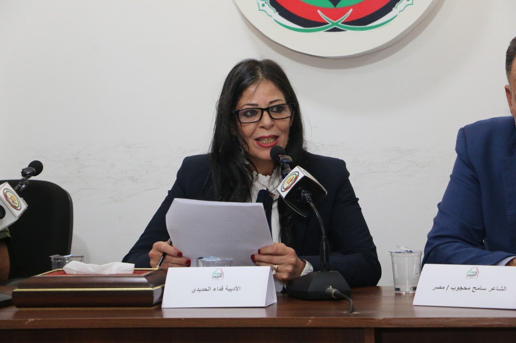 WhatsApp Image 2026-08-04 at 111736 AM1 Woman in a dark blazer and glasses speaking into a microphone at a panel, holding papers with a nameplate featuring Arabic text in front of her.