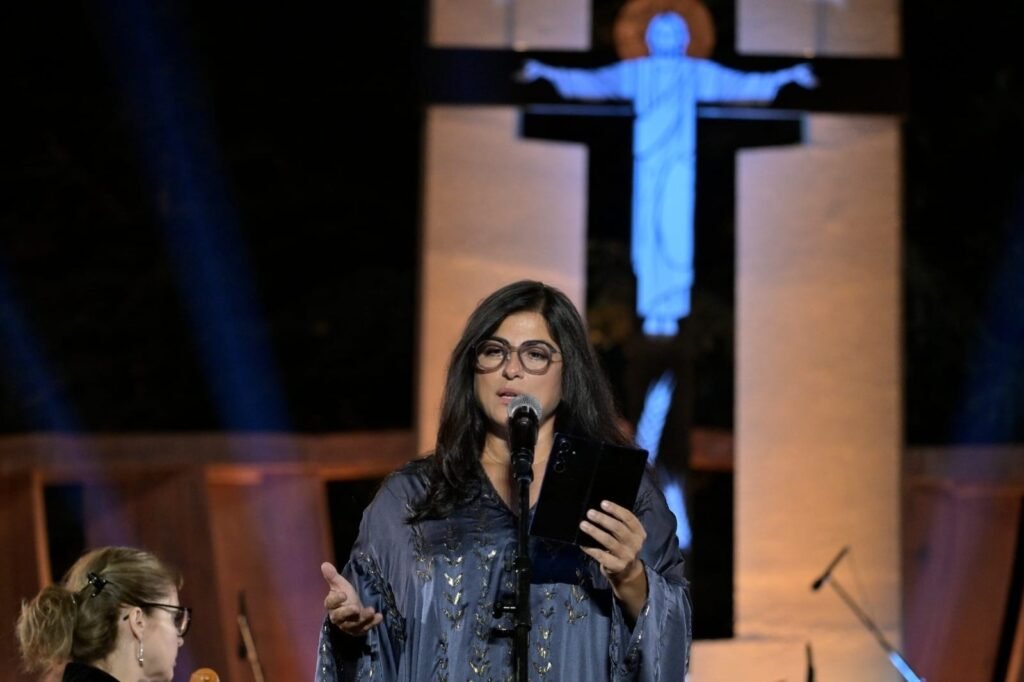 WhatsApp Image 2026-08-04 at 41235 PM - عروبة الاخباري | Oroba News A woman with glasses singing into a microphone on stage, holding a tablet in her left hand against a lit backdrop with a cross-like structure behind her.
