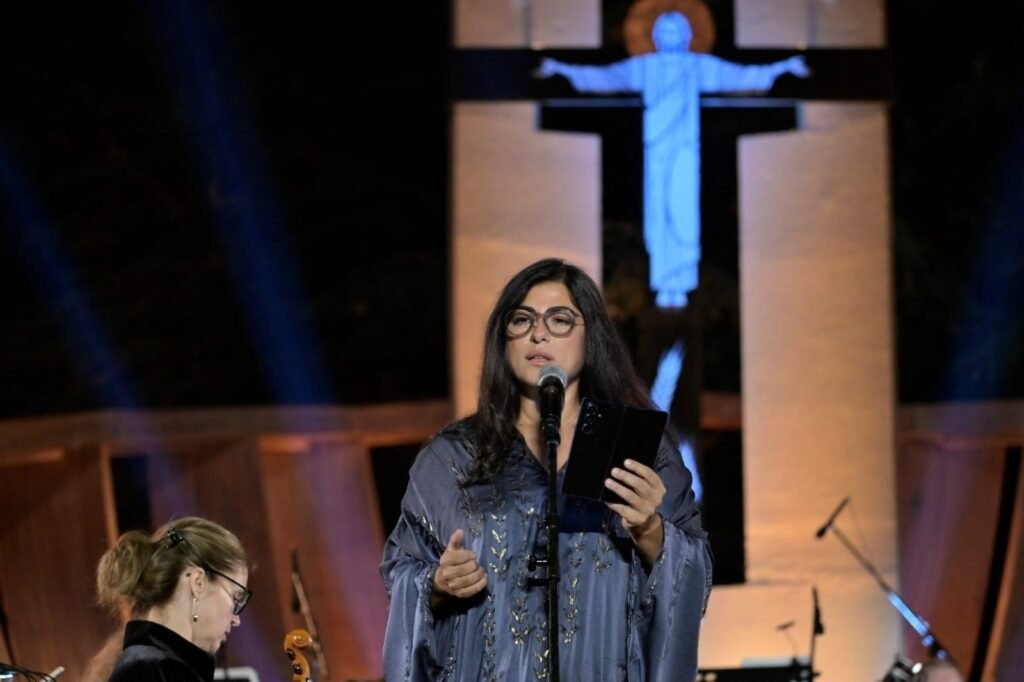 WhatsApp Image 2026-08-04 at 41235 PM 2 - عروبة الاخباري | Oroba News Female singer with glasses on stage, wearing a blue embroidered robe, holding a tablet and microphone, with a blue-lit crucifix behind her.