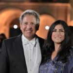 WhatsApp Image 2026-08-04 at 41235 PM 3 - عروبة الاخباري | Oroba News Smiling man in a black tuxedo with a white bow tie stand beside a woman in a blue embroidered dress at a formal evening event, arched warm-lit background behind them.