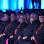 WhatsApp Image 2026-08-04 at 41235 PM 4 - عروبة الاخباري | Oroba News Row of clergy in black robes with red sashes seated in an audience at a formal event under blue lighting.