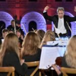 WhatsApp Image 2026-08-04 at 41236 PM - عروبة الاخباري | Oroba News Conductor leads a live orchestra in a dimly lit hall with red and blue stone arches in the background.