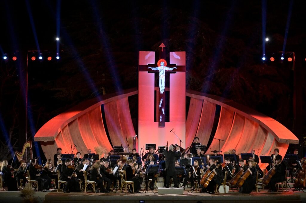 WhatsApp Image 2026-08-04 at 41236 PM 2 - عروبة الاخباري | Oroba News Outdoor orchestra concert at night on a curved orange-lit stage with a crucifix backdrop and conductor leading the ensemble.