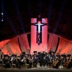 WhatsApp Image 2026-08-04 at 41236 PM 2 - عروبة الاخباري | Oroba News Nighttime outdoor orchestra on a lit stage, conductor at front, with a large cross sculpture and curved red-lit wooden backdrop behind them.