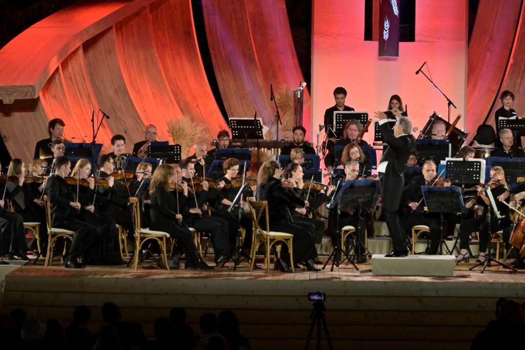 WhatsApp Image 2026-08-04 at 41236 PM 3 - عروبة الاخباري | Oroba News Full orchestra performing on stage with a conductor in front, warm orange lighting in the background, string players in the foreground.