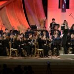 WhatsApp Image 2026-08-04 at 41236 PM 3 - عروبة الاخباري | Oroba News Full orchestra on stage with conductor leading, musicians reading sheet music in black formal attire, warm red lighting.